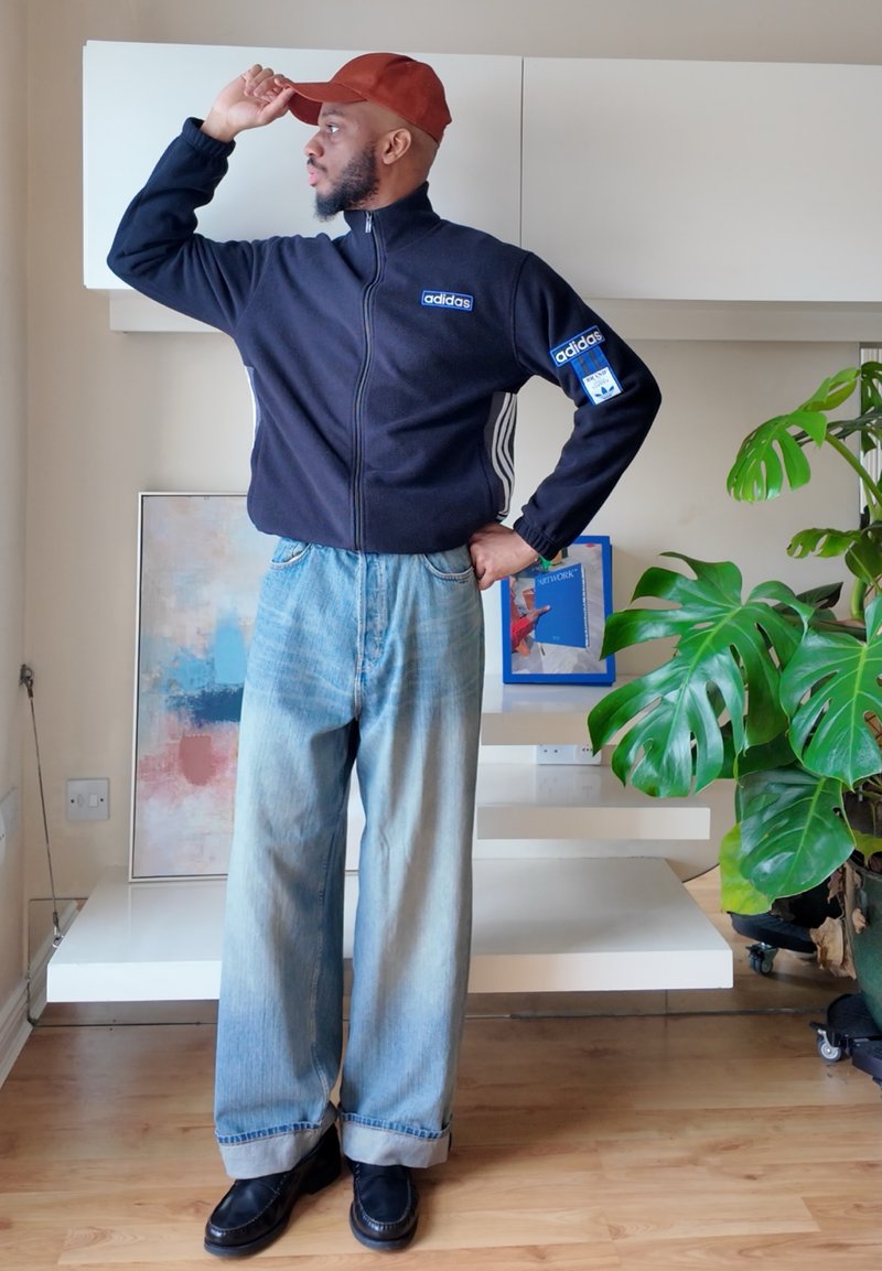 Homme portant une veste Adidas noire, un jean bleu clair à jambes larges, des chaussures noires et une casquette rouge, faisant un salut avec la main, debout à l'intérieur près d'une plante verte et d'une œuvre d'art.