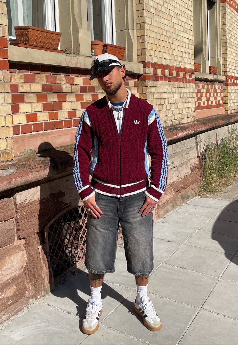 Homme portant une casquette blanche, une veste Adidas bordeaux avec des rayures bleues, un short gris, des chaussettes blanches et des baskets, debout sur le trottoir près d'un bâtiment en briques.