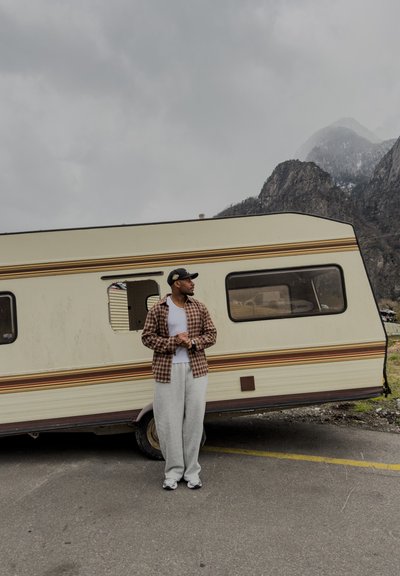Hombre vestido con ropa informal y gorra, de pie frente a una caravana beige con montañas y cielo nublado al fondo.
