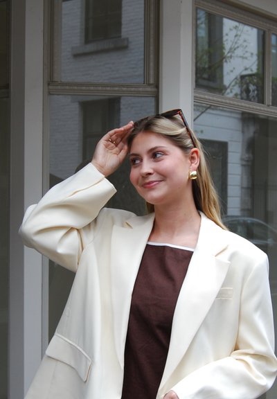 Blazer crema sobre una blusa marrón, pendientes dorados brillantes y gafas de sol rectangulares. El blazer tiene hombros estructurados y un bolsillo.