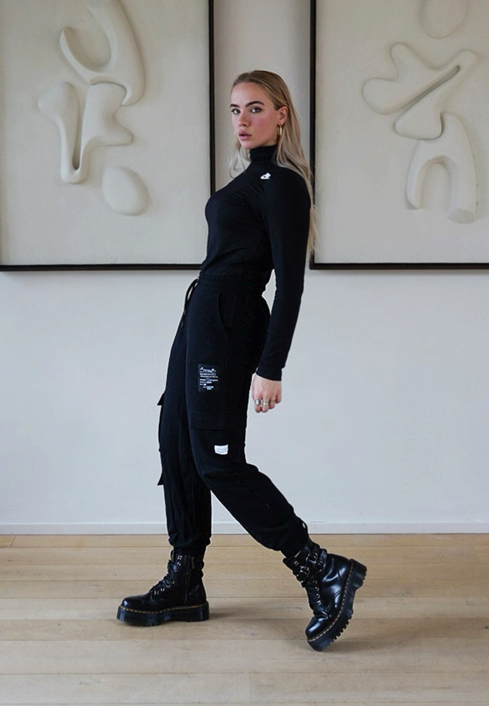 Camisola preta de mangas longas combinada com calças cargo pretas com bolsos de remendo. Botas de combate pretas com sola grossa. Fundo interior minimalista.
