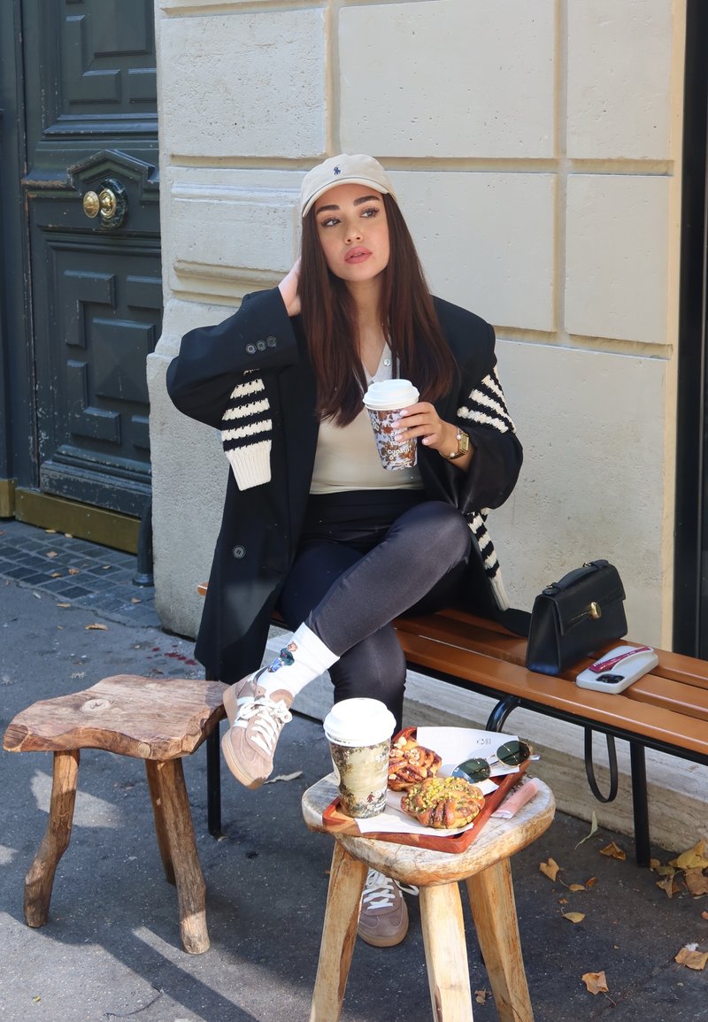 Blazer nero con strisce bianche, cappello beige e top crema. Seduta su una panchina di legno, tiene in mano una tazza di caffè ed è circondata da snack.