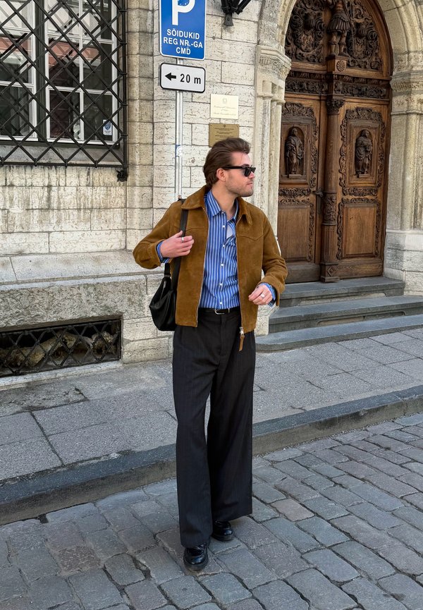 Bărbat purtând ochelari de soare, jachetă maro, cămașă albastră cu dungi și pantaloni negri stă lângă intrarea unei clădiri din piatră cu uși din lemn sculptat și semn de parcare.