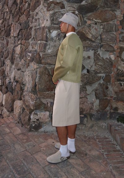 Suéter de punto verde claro, falda beige hasta la rodilla, calcetas blancas y zapatos grises sin cordones. Estilizado contra un fondo de pared de piedra.