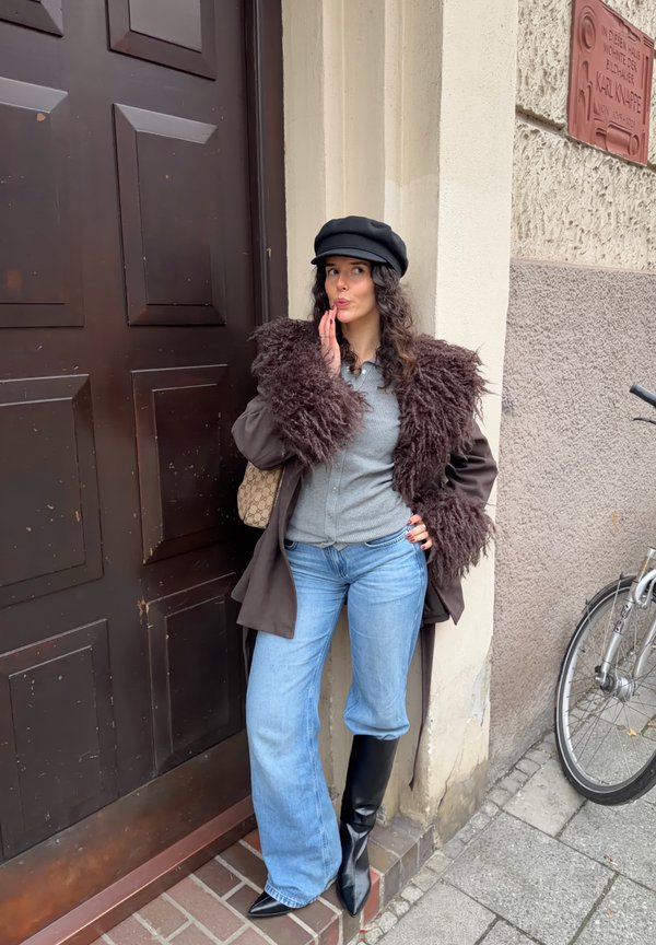 Brązowy płaszcz z puszystymi futrzanymi rękawami, szary top, jasnoniebieskie dżinsy i czarne kozaki za kolano; stoi przy brązowych drzwiach z ceglaną ścieżką.