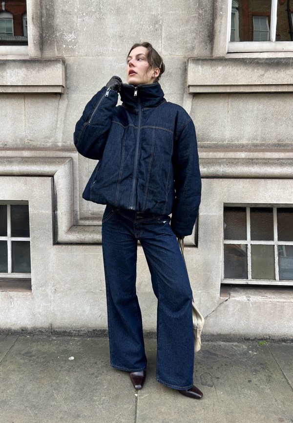 Blå denim puffjacka med hög krage och mörka vida jeans. Outfiten har kontrasterande sömmar och bruna stövlar med klack.