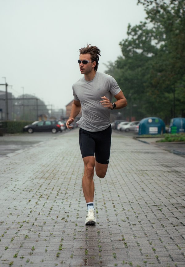 Un hombre con gafas de sol, una camiseta gris y pantalones cortos negros corre por un camino pavimentado cerca de coches aparcados y árboles.