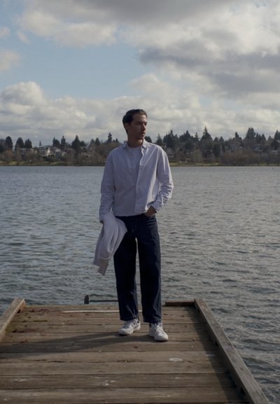 Hombre con ropa casual sosteniendo una chaqueta blanca, de pie en un muelle de madera junto a un lago tranquilo con casas y árboles en el fondo.