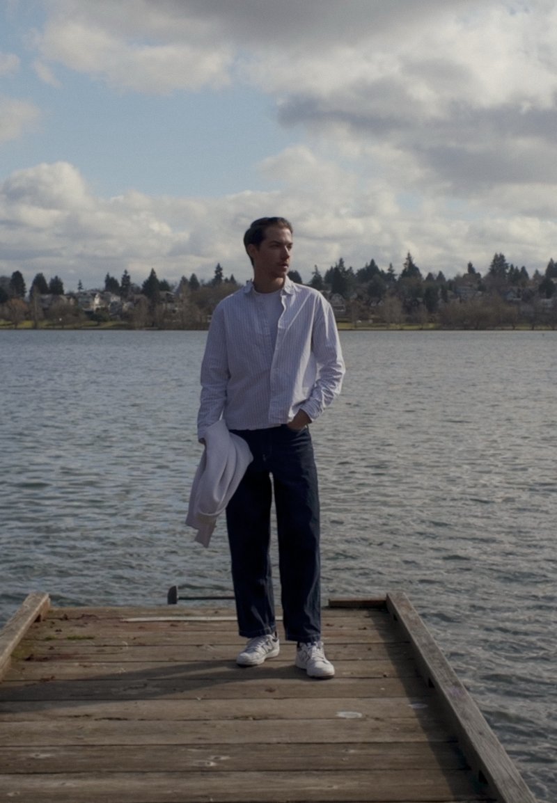 Mann in Freizeitkleidung hält eine weiße Jacke und steht auf einem Holzsteg an einem ruhigen See mit Häusern und Bäumen im Hintergrund.