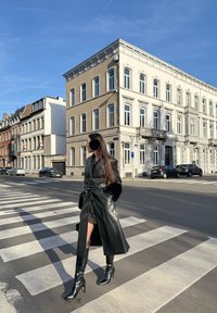 Hosszú fekete bőrkabát prémrészletekkel, térd fölé érő fekete magassarkú csizma és egy ruha. Fényteli napsütés és gyalogátkelő egy városi környezetben.