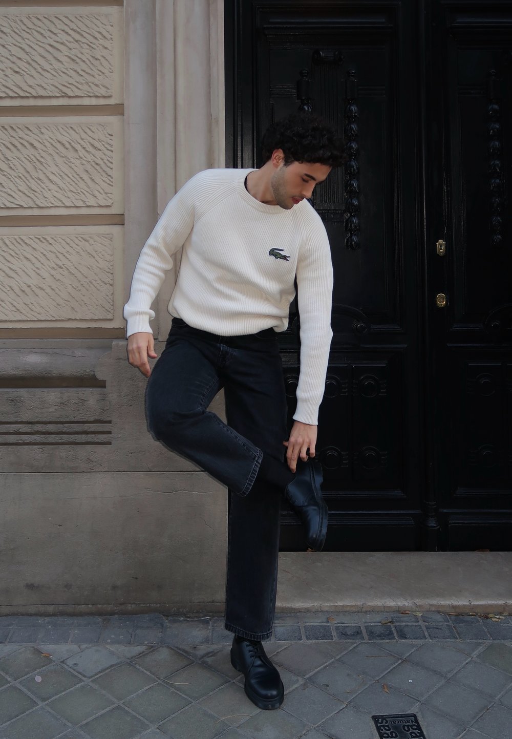 Λευκό ριγέ πουλόβερ με μικρό λογότυπο, μαύρα τζιν και μαύρα παπούτσια. Το μοντέλο ρυθμίζει ένα παπούτσι ενώ στέκεται σε πλακόστρωτο δρόμο.