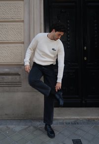 Λευκό ριγέ πουλόβερ με μικρό λογότυπο, μαύρα τζιν και μαύρα παπούτσια. Το μοντέλο ρυθμίζει ένα παπούτσι ενώ στέκεται σε πλακόστρωτο δρόμο.