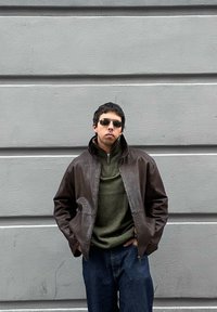 Homme portant des lunettes de soleil noires, une veste en cuir marron, un pull vert et un jean bleu, debout les mains dans les poches contre un mur gris en arrière-plan.