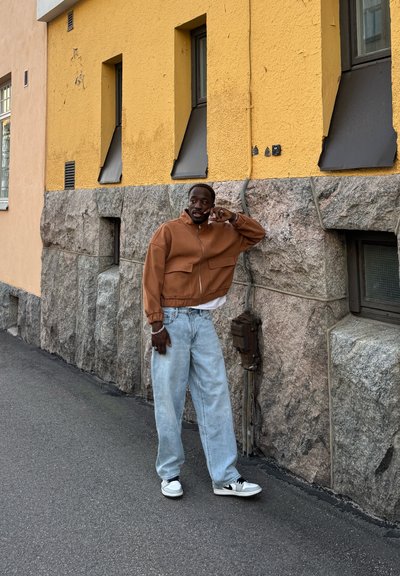 Chaqueta marrón corta con dos bolsillos frontales, vaqueros holgados de color azul claro y zapatillas negras y blancas, de pie contra una pared de piedra.