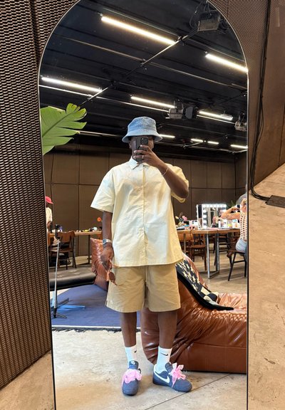 Camisa de botones de manga corta, amarilla clara; pantalones cortos beige; sombrero de cubo azul; zapatillas grises con acentos rosas; de pie frente a un espejo.