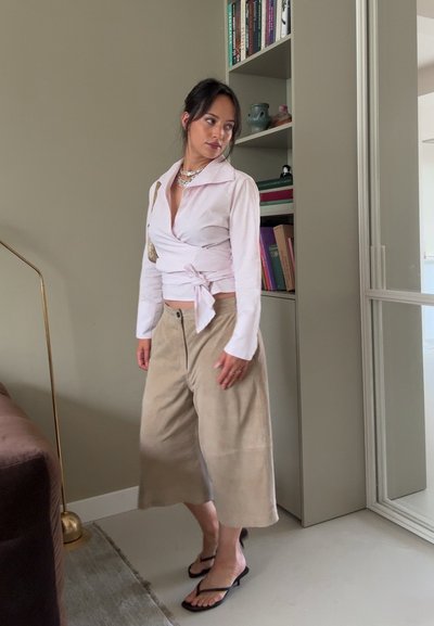 Blusa envolvente rosa claro con mangas largas y lazo en la parte delantera, combinada con pantalones de ante beige de pierna ancha y chanclas negras. Se pueden ver dos collares.