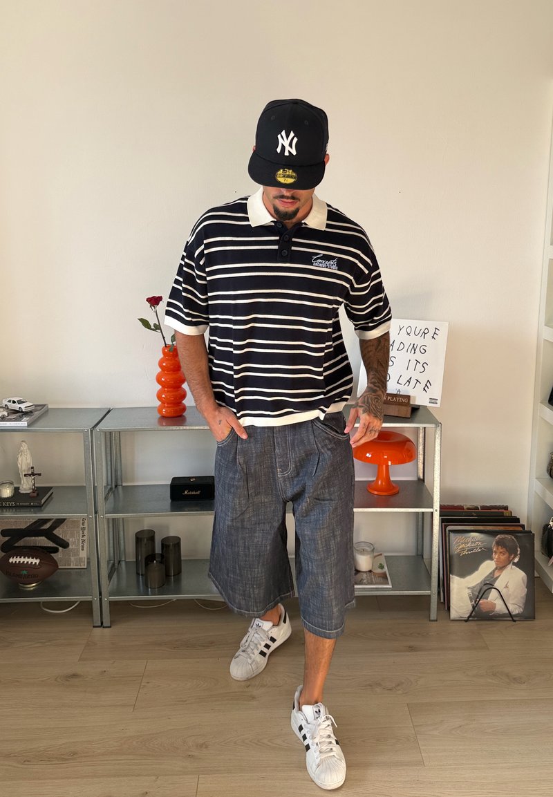 Homme portant un polo rayé, un short en jean, des baskets blanches et une casquette noire, debout à l'intérieur près d'étagères avec décoration et disques vinyles.
