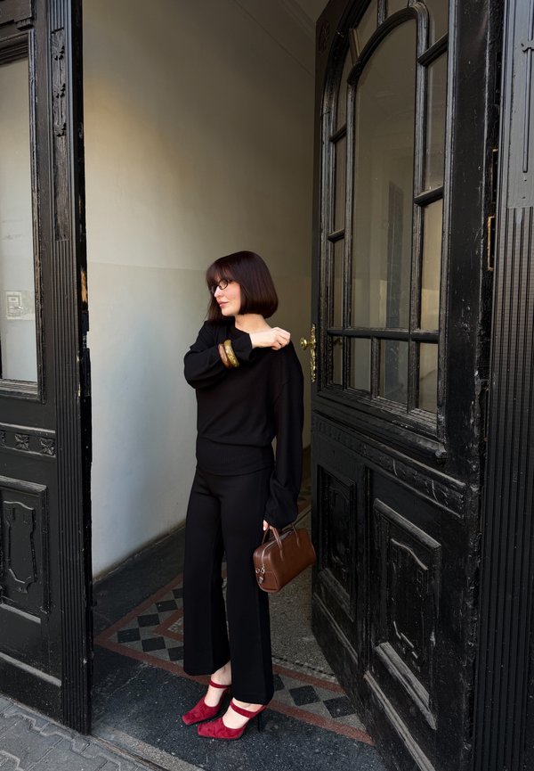 Lyhyttukkaisen ruskeahiuksisen naisen mustassa asussa ja punaisissa koroissa seisoo ja pitää ruskeaa käsilaukkua, säätäen hihaa avoimen mustan oven kohdalla.