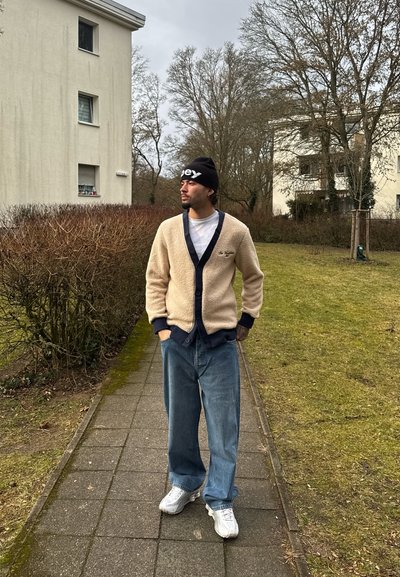 Cárdigan beige de peluche con cuello y botones azul marino, combinado con vaqueros azules holgados y zapatillas blancas, llevado con un gorro negro.