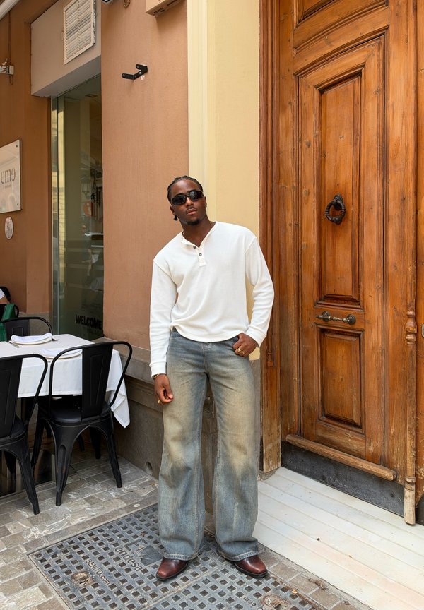 Un hombre con gafas de sol, una camisa blanca y vaqueros holgados está junto a una puerta de madera tallada fuera de un restaurante.