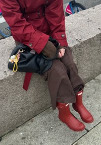 Røde gummistøvler med hvit logo, kombinert med brune bukser med tynne striper og en rød jakke, hvilende på en steinoverflate. Svart veske med nøkkelring.
