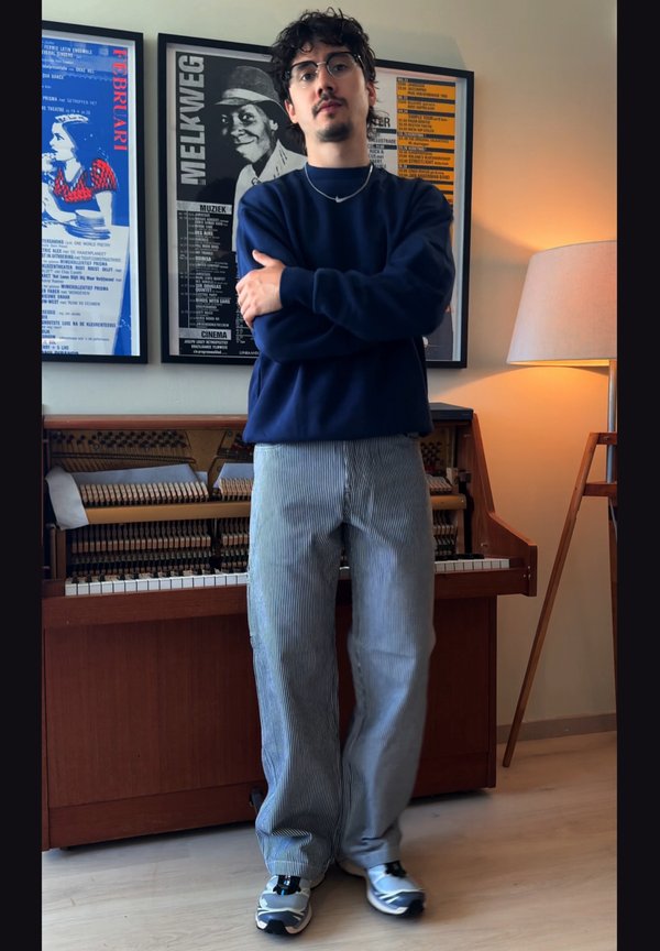 Hombre con gafas, suéter azul y pantalones a rayas está de pie con los brazos cruzados frente a un piano abierto y carteles enmarcados en la pared.