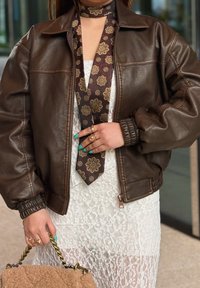 Bruine leren bomberjack met ribbels aan de manchetten, gecombineerd met een gestreepte bruin sjaal. Witte kanten jurk van een lichte, gemêleerde stof.