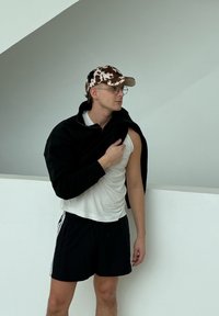 Νέος άνδρας που φοράει καπέλο με μοτίβο αγελάδας, γυαλιά, λευκό αμάνικο, μαύρο σορτς και μαύρο μπουφάν περασμένο πάνω σε έναν ώμο, μπροστά από έναν απλό τοίχο.