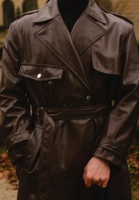 Abrigo trench coat de cuero marrón con solapas grandes, botones de doble hilera, bolsillos laterales y cinturón en la cintura. Textura suave.