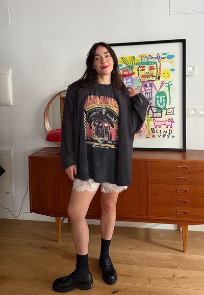 Mujer con cabello largo y oscuro que lleva una blazer negra, camiseta gráfica, pantalones cortos blancos de encaje, calcetines y zapatos negros, de pie frente a una cómoda de madera y una obra de arte colorida.