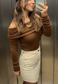 Hnědý top s odhalenými rameny spárovaný s bílou džínovou sukní. Významné doplňky zahrnují vrstevnatý náhrdelník a prsteny. Jednoduché pozadí.