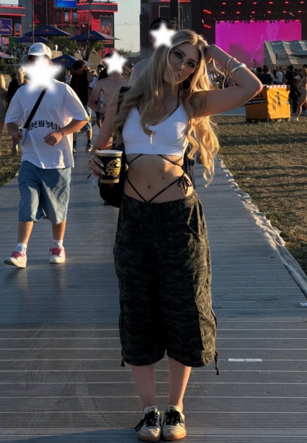 Jeune femme aux longs cheveux blonds portant un haut blanc et un pantalon camouflage, tenant une boisson, debout sur un chemin en bois lors d'un événement en plein air.