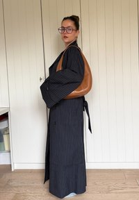 Čierny dlhý kabát s tenkými pásikmi, veľkými rukávmi a bočným zaväzovaním. Hnedá kožená kabelka na rameno. Svetlá drevená podlaha a jednoduché pozadie.