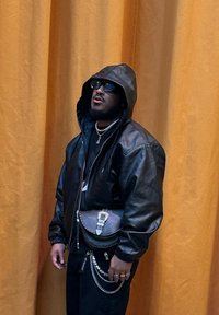 Veste en cuir noire avec capuche, pantalon noir et un sac à dos rond. Accentué de chaînes et d'anneaux en argent. Fond en tissu orange.