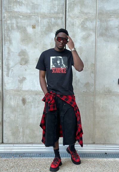 Camiseta negra con gráfico de "Scarface", combinada con pantalones cortos negros desgastados. Camisa de cuadros roja atada a la cintura, zapatillas rojas y negras.