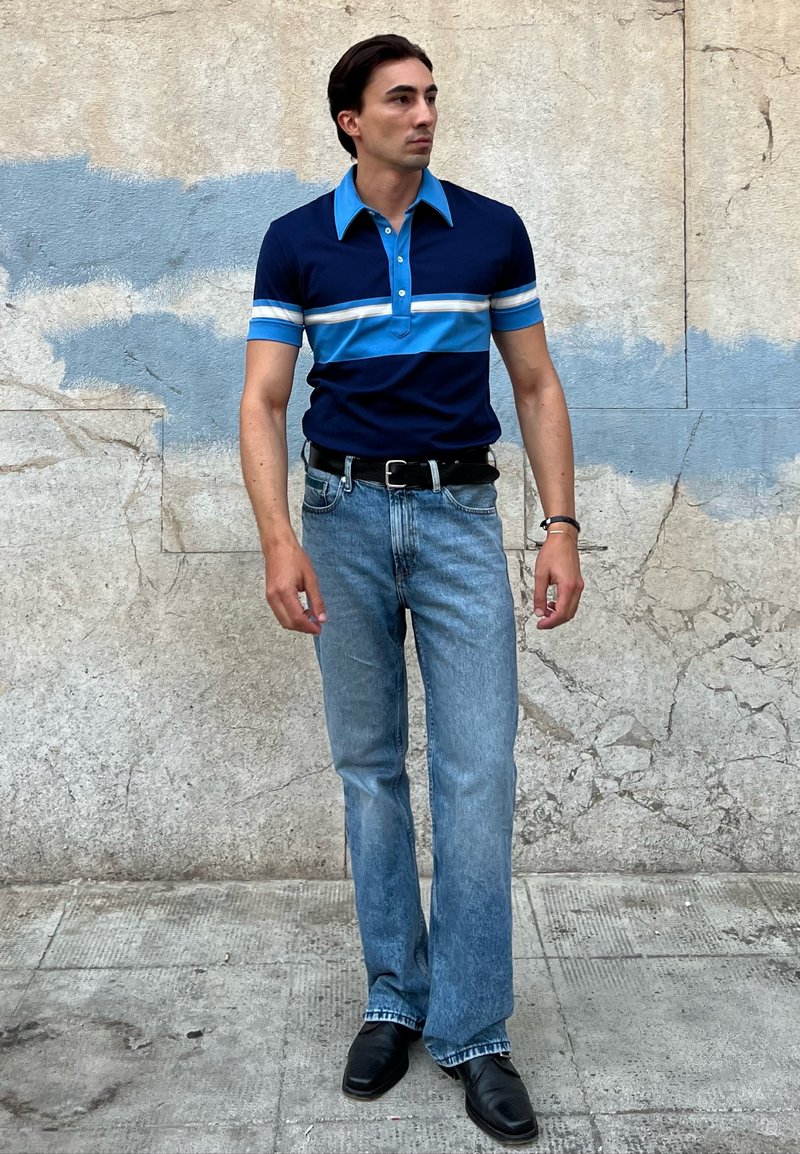 Pólo listrado em azul-marinho e azul-claro com mangas curtas, combinado com jeans claros desbotados e sapatos sociais pretos. Cor sólida e corte clássico.