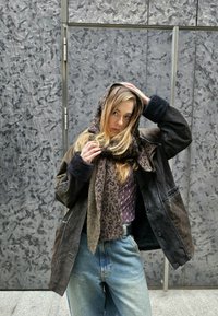 Καφέ δερμάτινο μπουφάν με μαύρους κουμπαράδες από γούνα, κασκόλ με σχέδιο λεοπάρ, ανοιχτό γαλάζιο τζιν παντελόνι, μπροστά σε υφή γκρι φόντο.