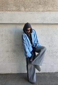 Personne portant une veste Adidas bleue, un pantalon large gris, un sweat à capuche noir, des lunettes de soleil et des gants, levant légèrement une jambe tout en se tenant contre un mur texturé.