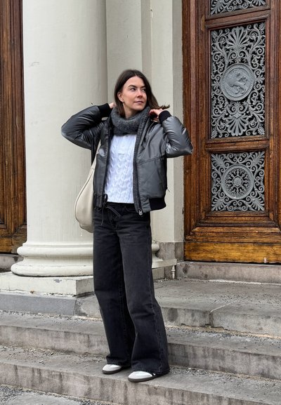 Chaqueta acolchada gris con cremallera, suéter de punto grueso color crema, jeans de pierna ancha negros y bufanda gris. De pie junto a una puerta de madera ornamentada en unas escaleras de piedra.