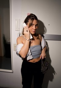 Bijela fiksna telefonska slušalica držana na uhu, u sivoj krojenoj majici i crnim hlačama, držeći malu tamnu torbicu, s svetlom pozadinom.