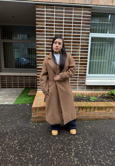 Abrigo de lana marrón con un collar ancho y mangas largas, combinado con una camiseta gris, pantalones oscuros y calzado marrón claro, de pie al aire libre.