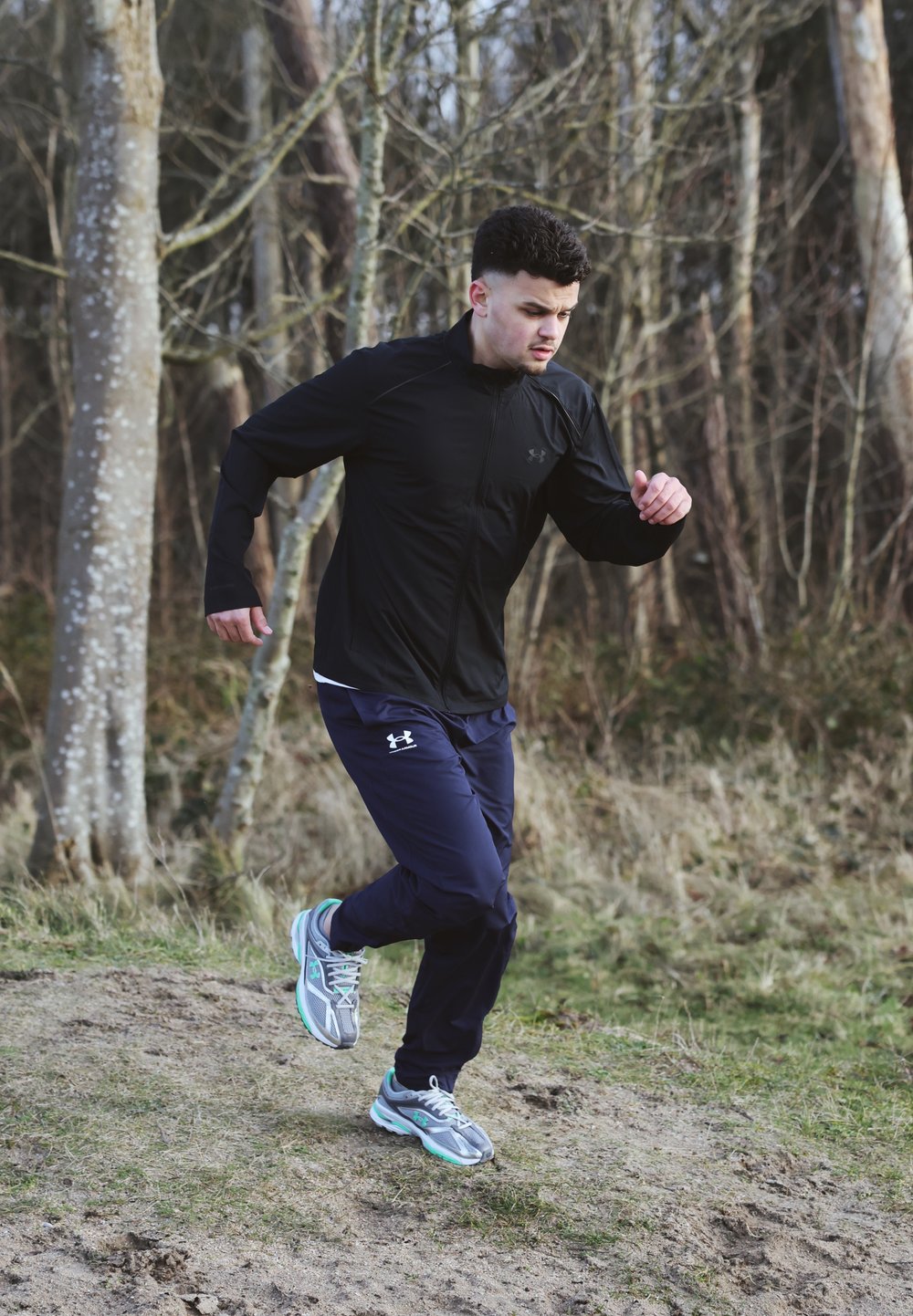 Young man in a black jacket and blue trousers jogging along a dirt path in a wooded area during the day.