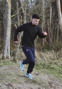 Young man in a black jacket and blue trousers jogging along a dirt path in a wooded area during the day.