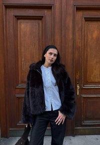 Jachetă din blană sintetică neagră cu glugă, bluză albă cu nasturi pe față și jeansi închiși la culoare, pe fondul unei pereți din lemn. Texturi și contraste notabile.