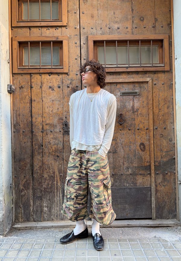 Jeune homme portant des lunettes de soleil, une chemise blanche à manches longues et un short camouflage se tient les mains dans les poches devant une grande porte en bois avec des fenêtres grillagées.