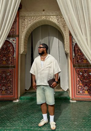 Camisa de manga corta color crema, pantalones cortos de mezclilla claros y zapatillas beige. De pie frente a un arco ornamentado y colorido con una cortina blanca fluida.