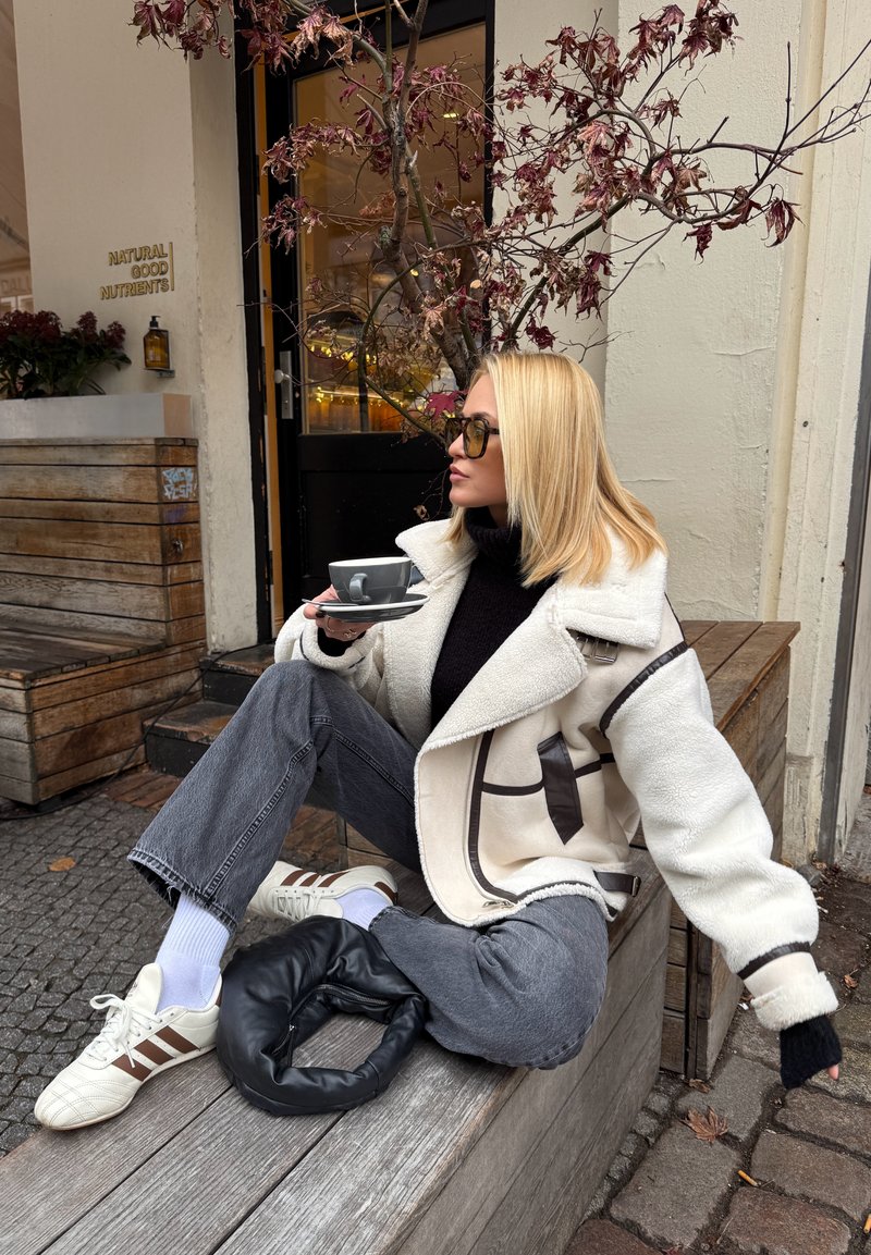 Casaco de lã com detalhes castanhos, gola alta preta, jeans cinzentos, sapatilhas brancas com riscas castanhas e uma mala preta circular. Chávena num pires.