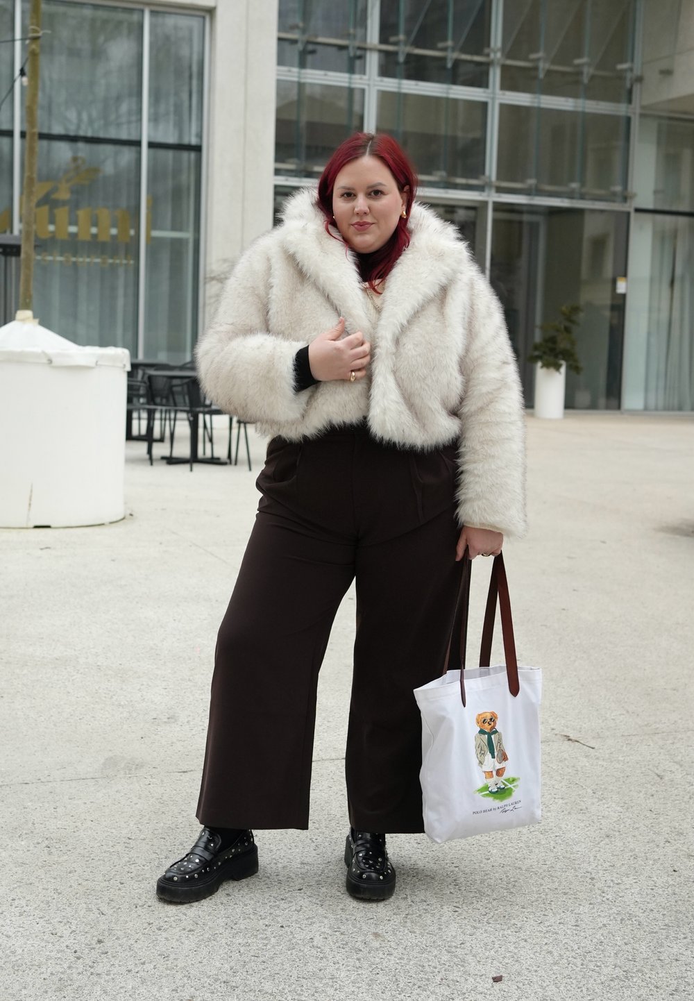 Femme aux cheveux roux portant une veste en fausse fourrure blanche, un pantalon noir et des chaussures noires avec des clous, tenant un sac fourre-tout blanc avec une illustration d'ours.