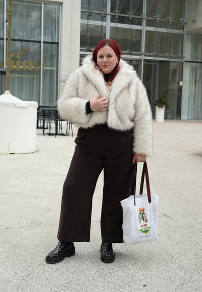 Mujer con cabello rojo que lleva una chaqueta blanca de piel sintética, pantalones negros y zapatos negros con tachuelas, sosteniendo un bolso tote blanco con una ilustración de un oso.