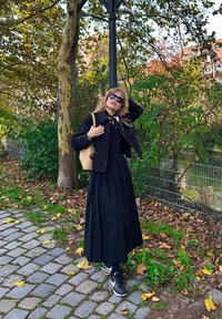 Jupe longue noire, haut noir ajusté, veste courte. Les accessoires incluent une écharpe à motifs et un sac à main de couleur claire. La mannequin porte des lunettes de soleil et des baskets.